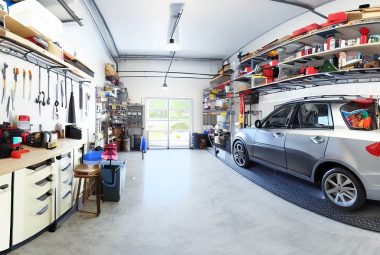 a panoramic view of a well-organized, clean, and bright garage. On the left, there is a car parked neatly. The walls show cool garage ideas with various storage systems like wall shelves, pegboards with tools, and cabinets. Overhead there are hanging storage bins and racks with items like camping gear or holiday decorations. On the right is a dedicated workspace with a sturdy workbench and a stool.