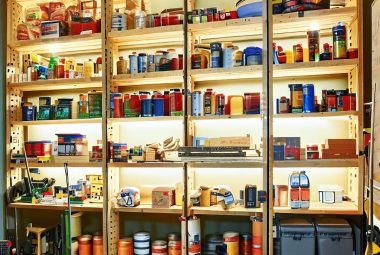 an image showcasing a well-organized, brightly lit garage space. In the middle of the image, include DIY wooden shelves made from 2x4's, filled with a variety of items like tools, paint cans, and storage boxes. The shelves should appear sturdy and fit well into the garage space, reaching almost up to the ceiling. The items on the shelves should be neatly arranged, demonstrating efficient use of space. Also, include a workbench in the scene with a tape measure, saw, and drils.