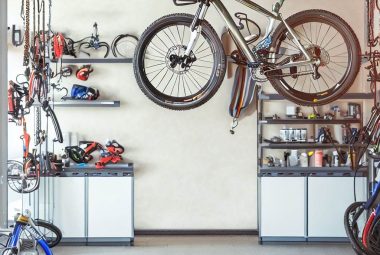 A tidy and organized garage interior showing multiple bike storage solutions. On one side of the garage, a bike is hung from a ceiling lift system, while another bike is hooked on a wall hanger. The other side features a free-standing bike rack holding two bikes and a single bike neatly placed on a floor stand. Tools and other garage items are organized on shelves and cabinets. The image is brightly lit, emphasizing the clean and clutter-free space.