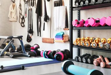 An image of a well-organized garage gym space with a mix of fitness equipment, such as dumbbells, a yoga mat, resistance bands, and a foldable workout bench. In the background, shelves with neatly arranged gym accessories and hooks holding exercise bands. The image clearly shows several home garage half gym ideas have been set up.