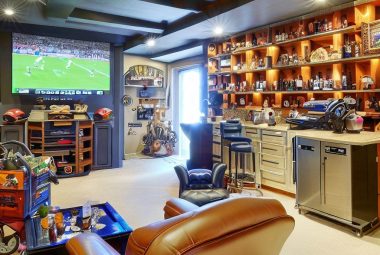 An image depicting a beautifully transformed garage into a man cave. The foreground features a comfortable recliner, a mini bar stocked with beverages, and a large screen TV displaying a sports game. The background showcases neatly organized wall-mounted racks holding tools, sports memorabilia, and other personal collections. Some overhead storage bins can be seen in the far end of the room.